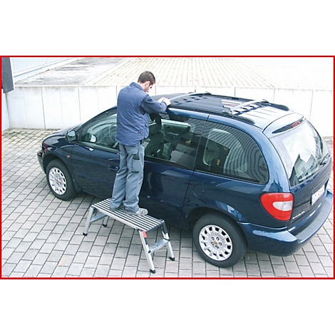 Ein Mann steht auf einer Trittleiter und arbeitet an einem blauen Van. Der Mann trägt eine Jacke und Jeans. Das Fahrzeug steht auf einer gepflasterten Fläche.